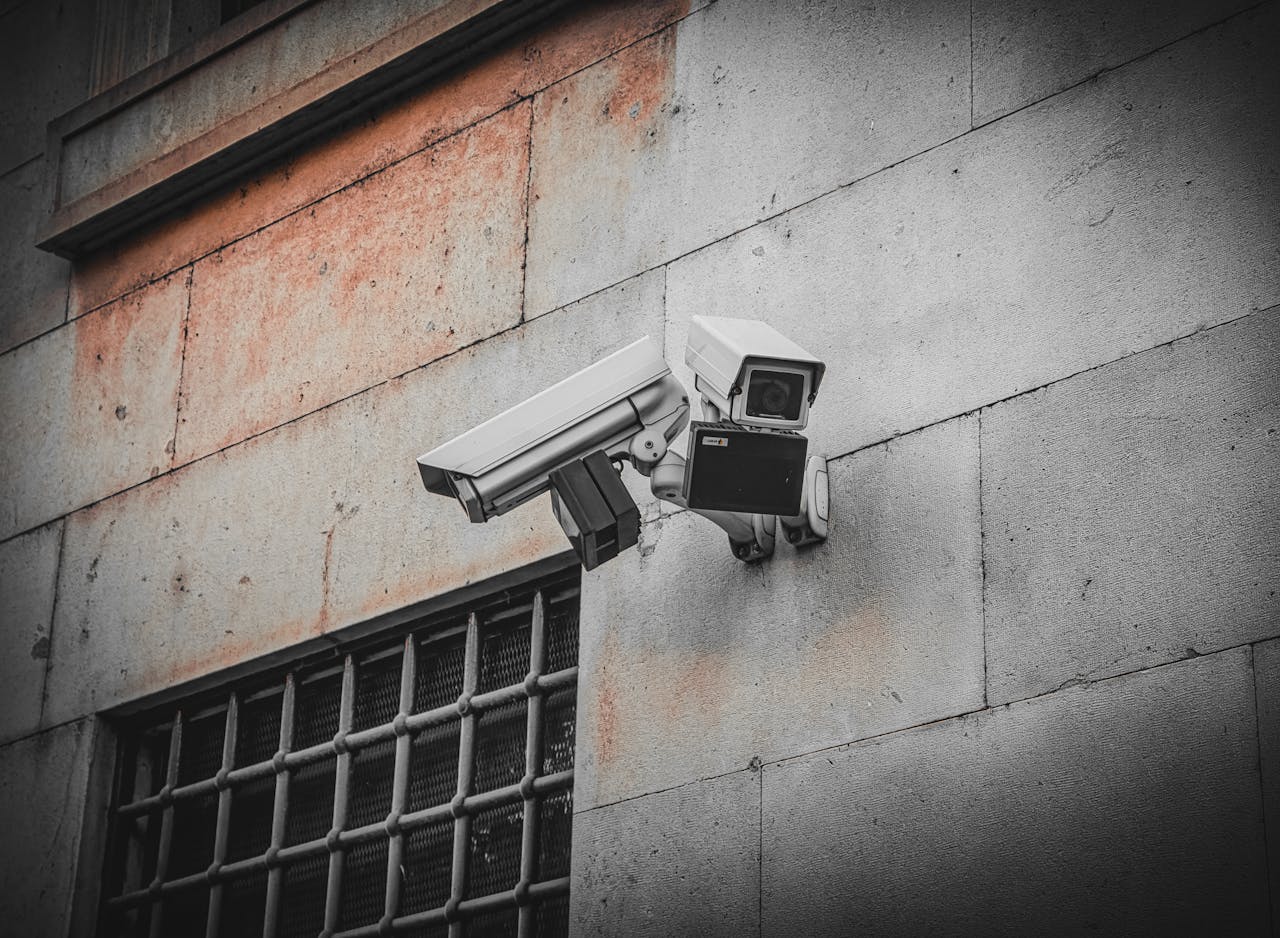 Two surveillance cameras mounted on an aged concrete wall beside a barred window, capturing urban security.
